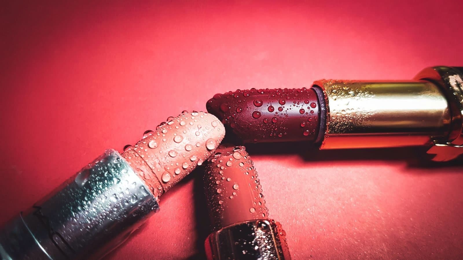 Close-up of colorful lipsticks with water droplets on a pink background, highlighting beauty.